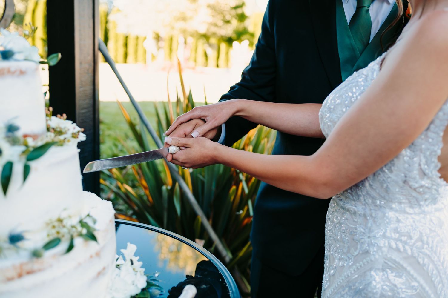 Bride and groom cutting their white wedding cake decorated with flowers and greenery at their reception.