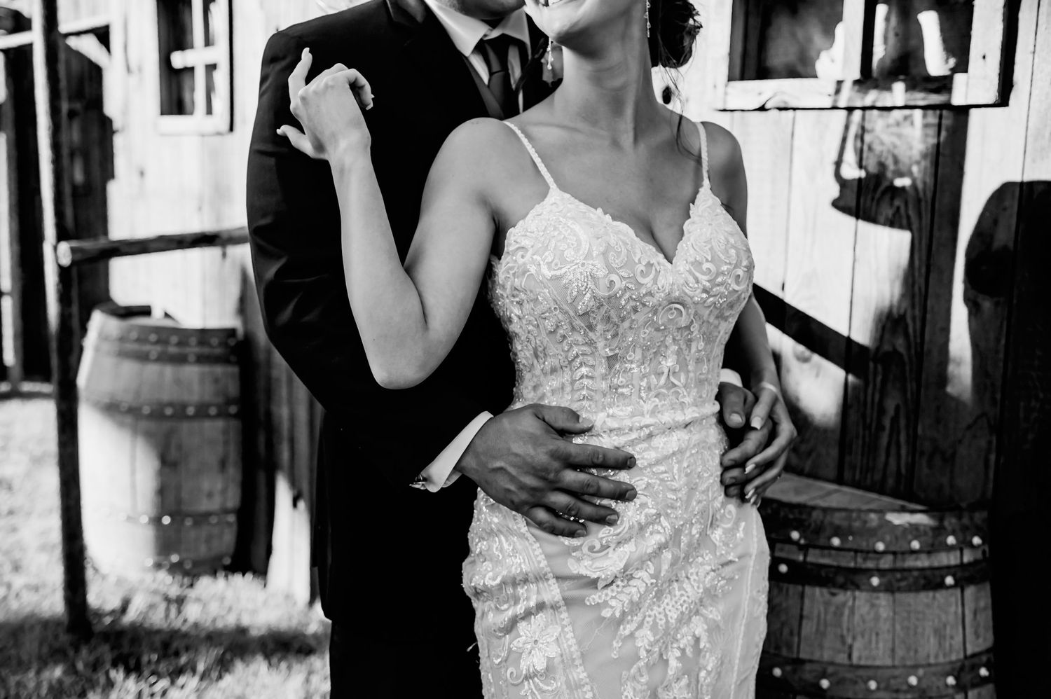 Black and white photo of a bride in a lace gown and groom in a suit embracing outdoors near rustic wooden barrels.