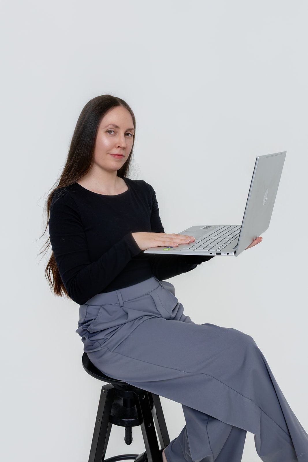 Smiling woman with long dark hair sitting on a stool, typing on a laptop, wearing black top and gray pants.