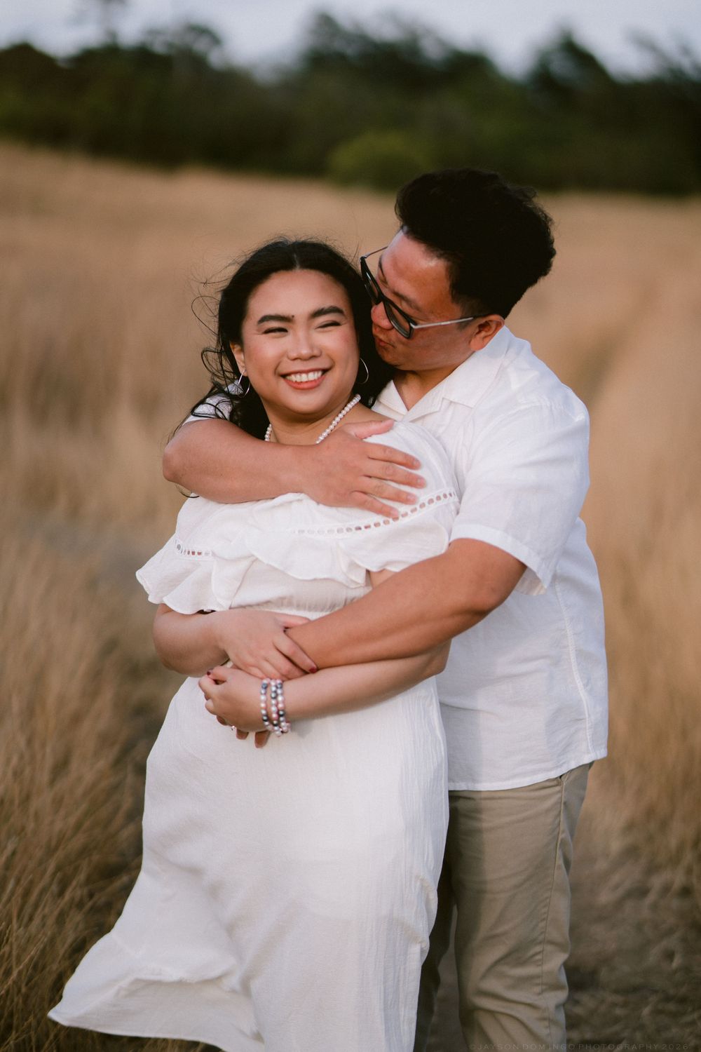 Couple embracing in golden field, woman in white off-shoulder dress smiling, man in white shirt kissing her cheek.