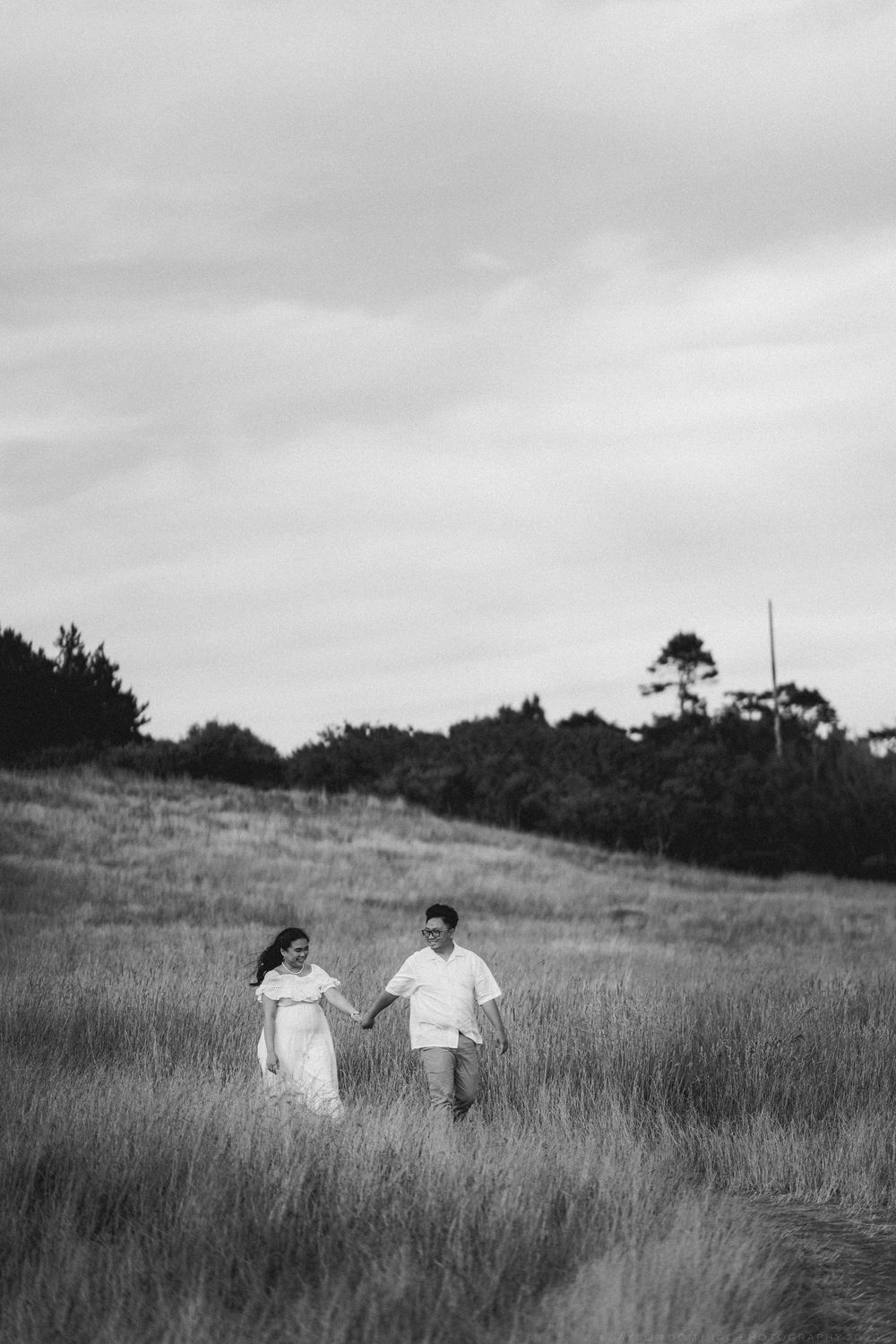 Black and white photo of a couple holding hands while walking through a grassy meadow with trees in the background.