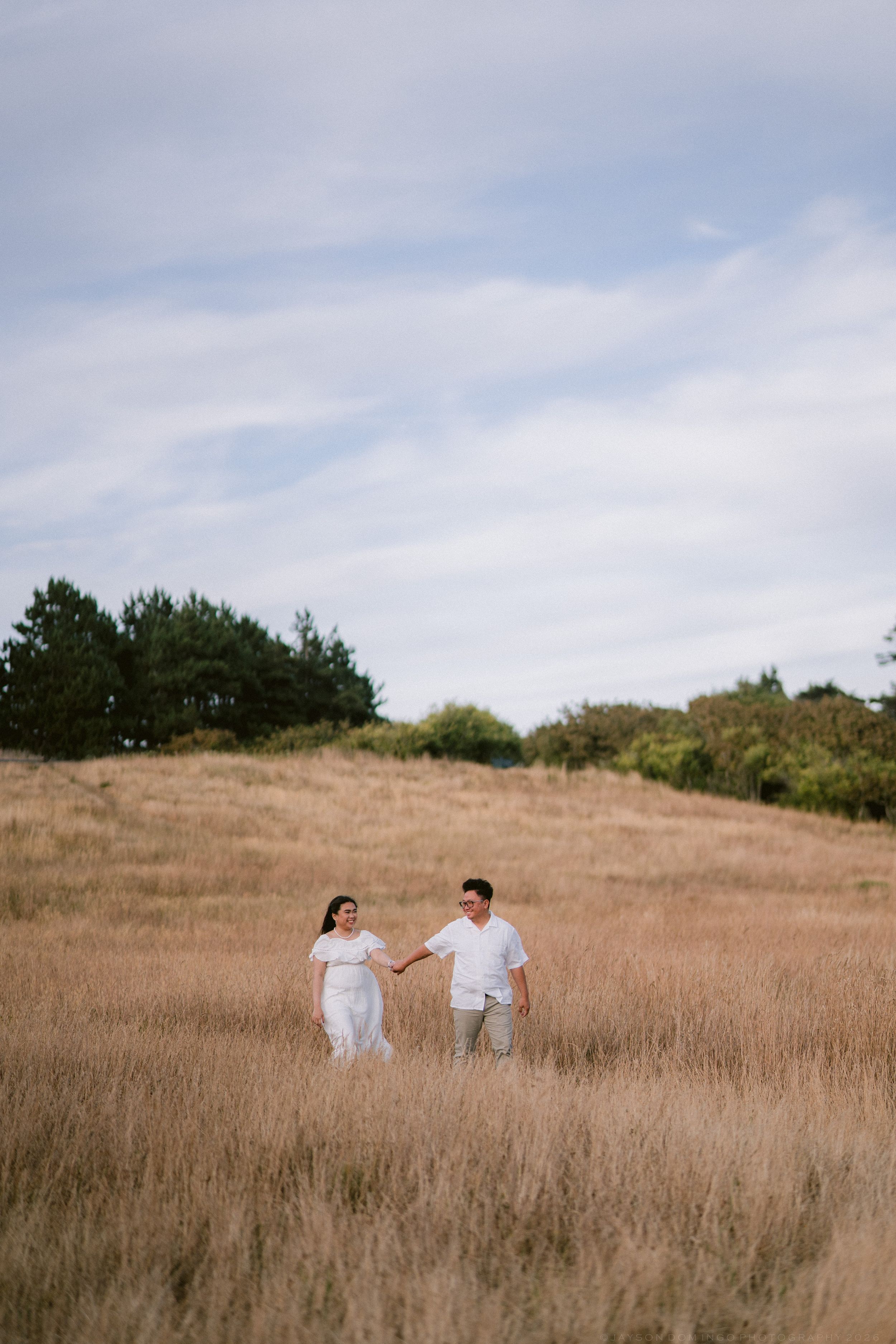 Couple holding hands in white outfits strolling through golden grass field with trees under cloudy sky.