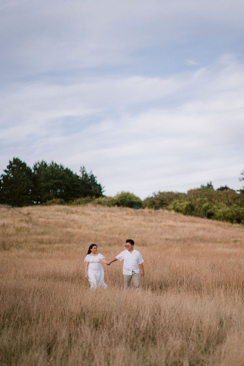 Couple holding hands in white outfits strolling through golden grass field with trees under cloudy sky.