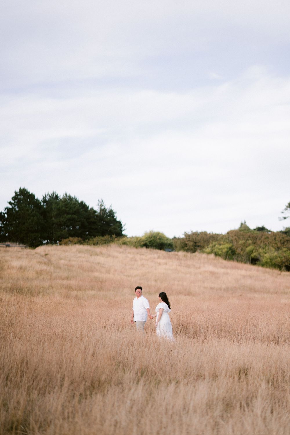 Couple in white outfits walking through golden grass field with trees in background.