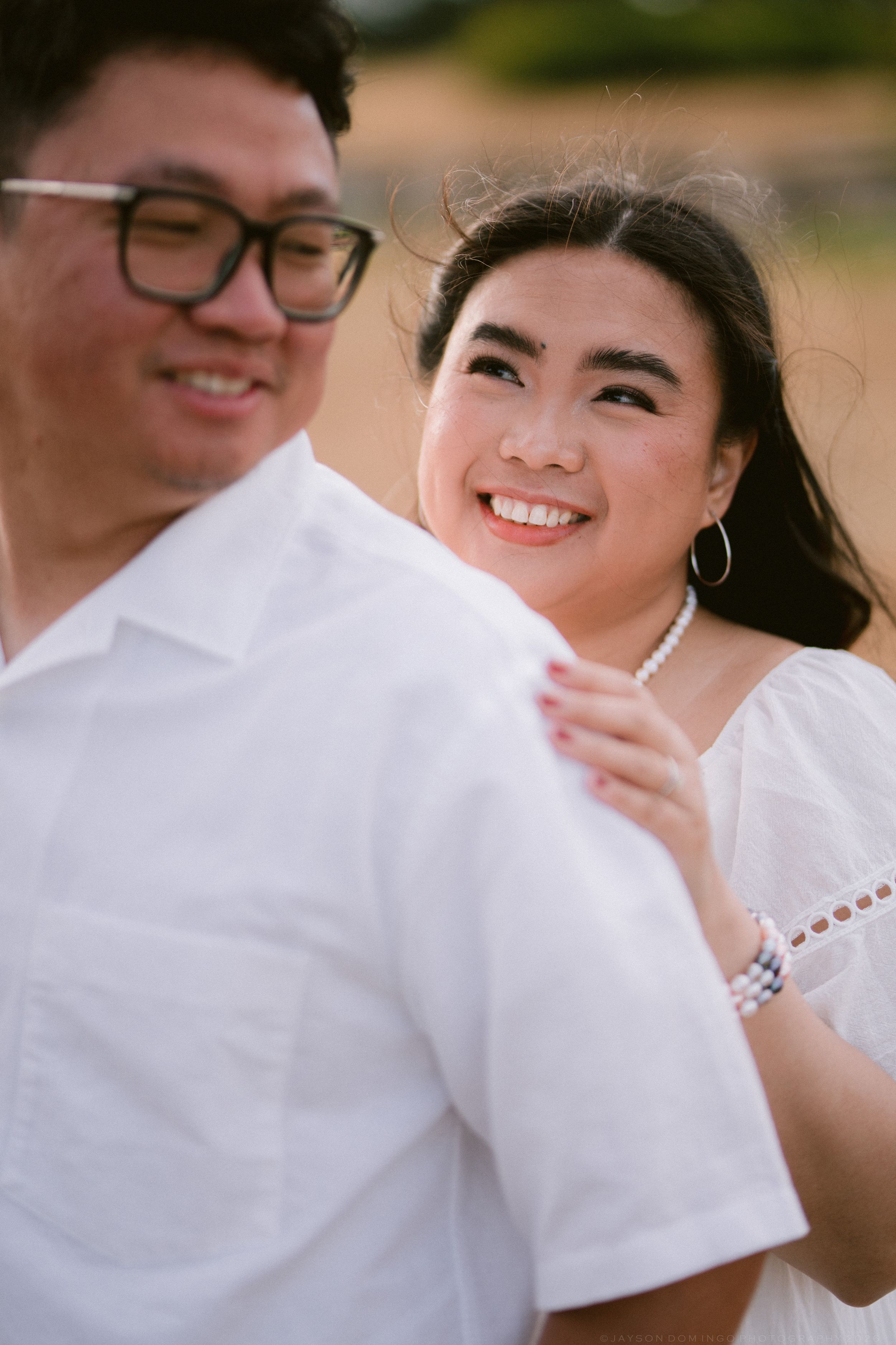 Woman in white dress smiling while embracing man with glasses outdoors.