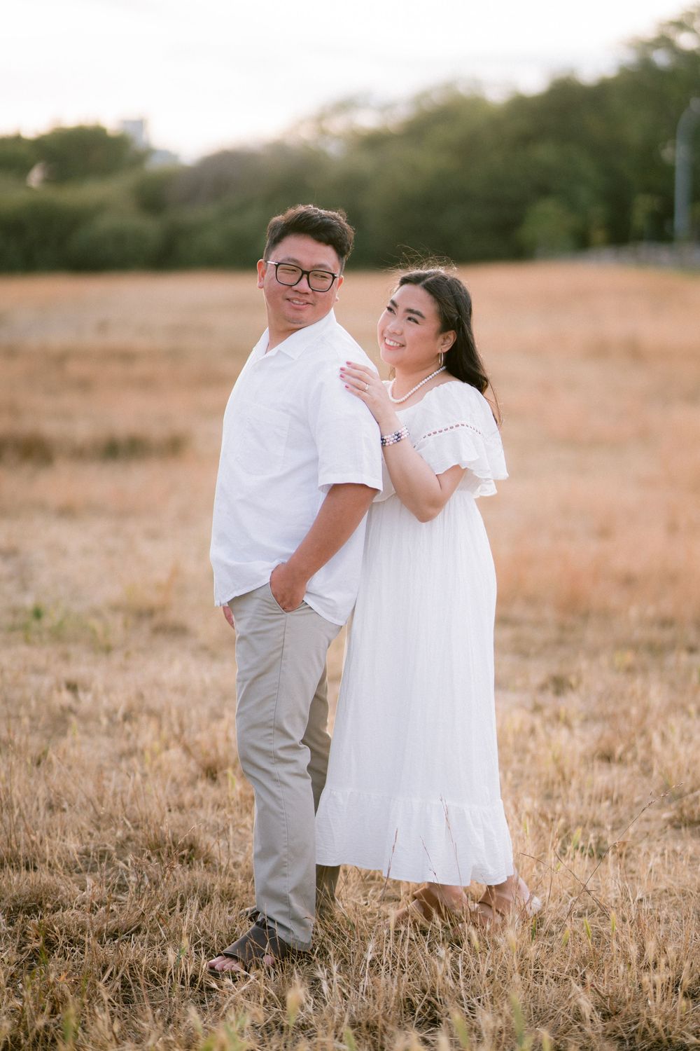 Couple in white outfits posing in golden field at sunset, woman in off-shoulder dress embracing man in glasses.