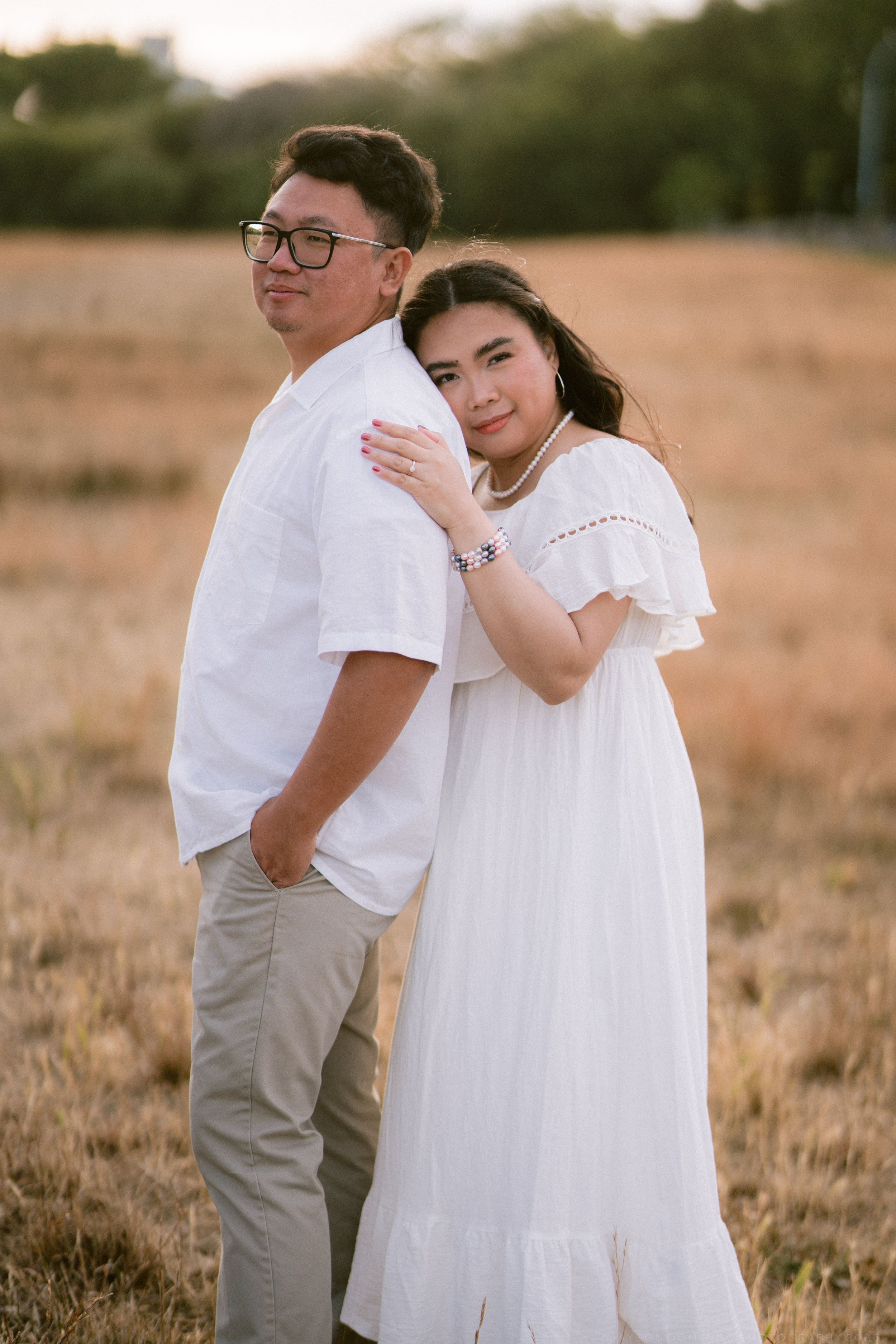 Couple posing in golden field, woman in white dress hugging man from behind.
