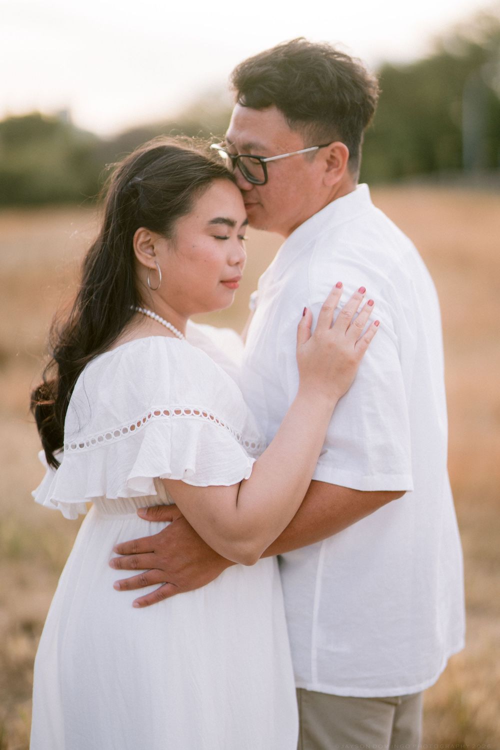 Asian couple in white embracing tenderly in golden field at sunset.