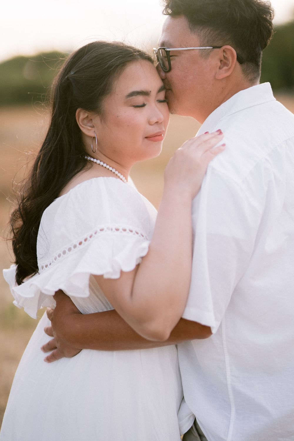 Asian couple embracing tenderly at golden hour, woman in white ruffled dress with pearl necklace, man kissing her forehead.