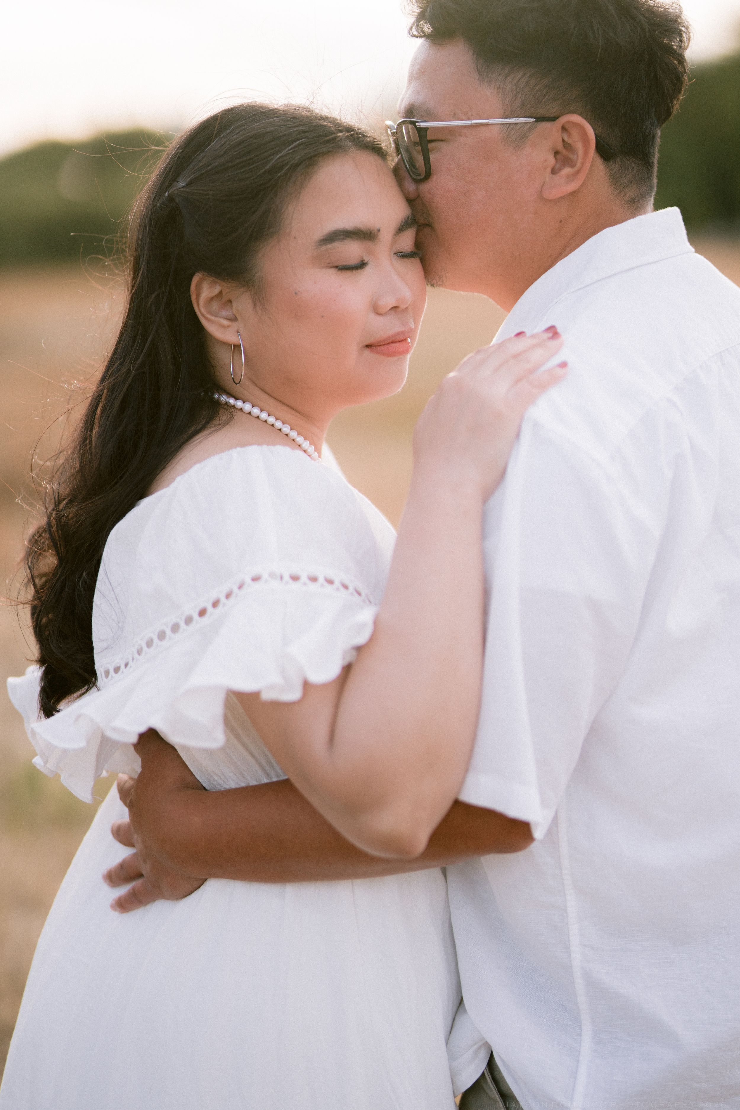 Asian couple embracing tenderly at golden hour, woman in white ruffled dress with pearl necklace, man kissing her forehead.