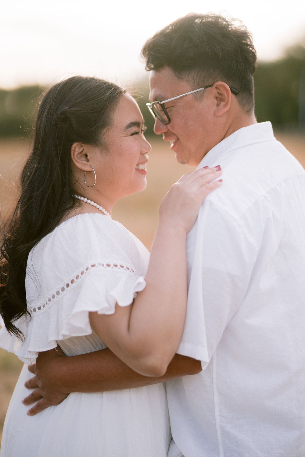 Happy couple in matching white outfits embrace and smile at each other during a romantic outdoor photo session.
