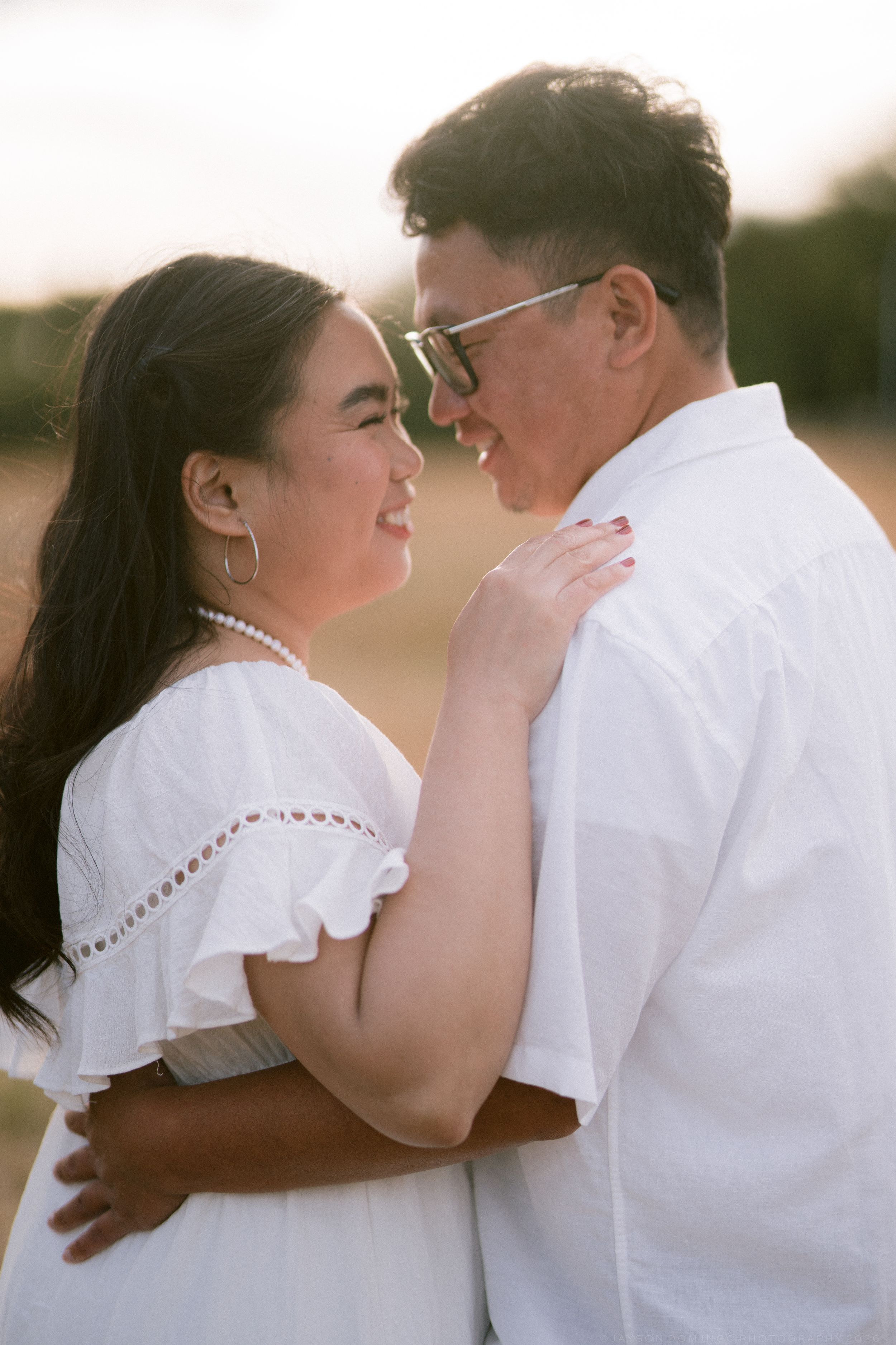 Couple in white outfits gazing at each other in sunny outdoor field setting.