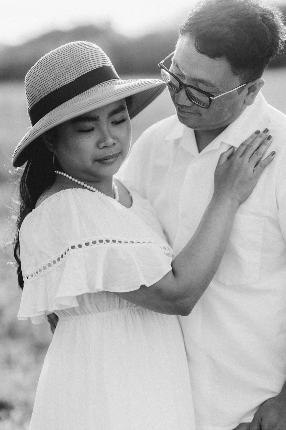 Black and white photo of a couple embracing outdoors, woman in white dress and sun hat, man in white shirt with glasses.