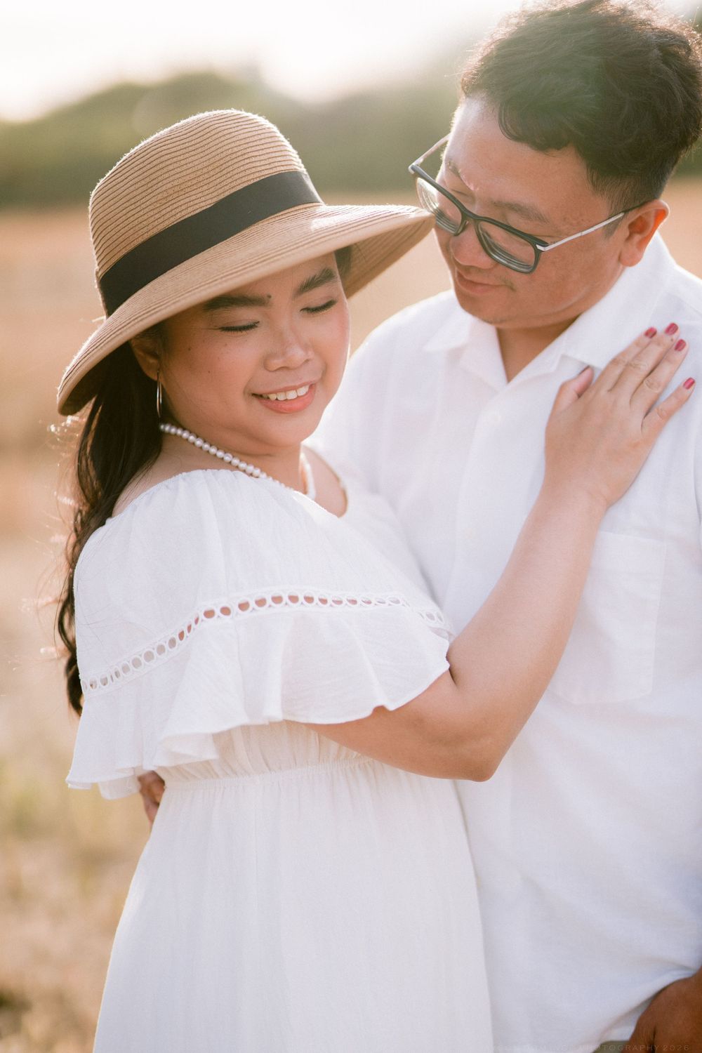 Couple in white outfits share a tender moment outdoors, woman wearing straw hat and pearl necklace at golden hour.