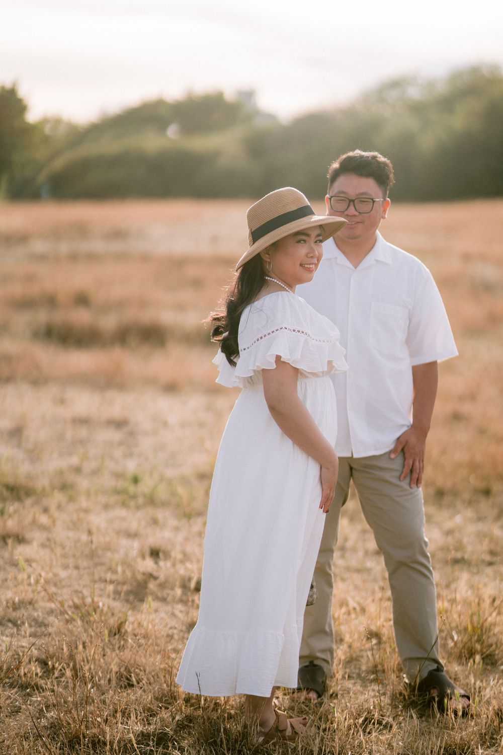 Smiling Asian woman in white dress and hat with partner in sunny field.