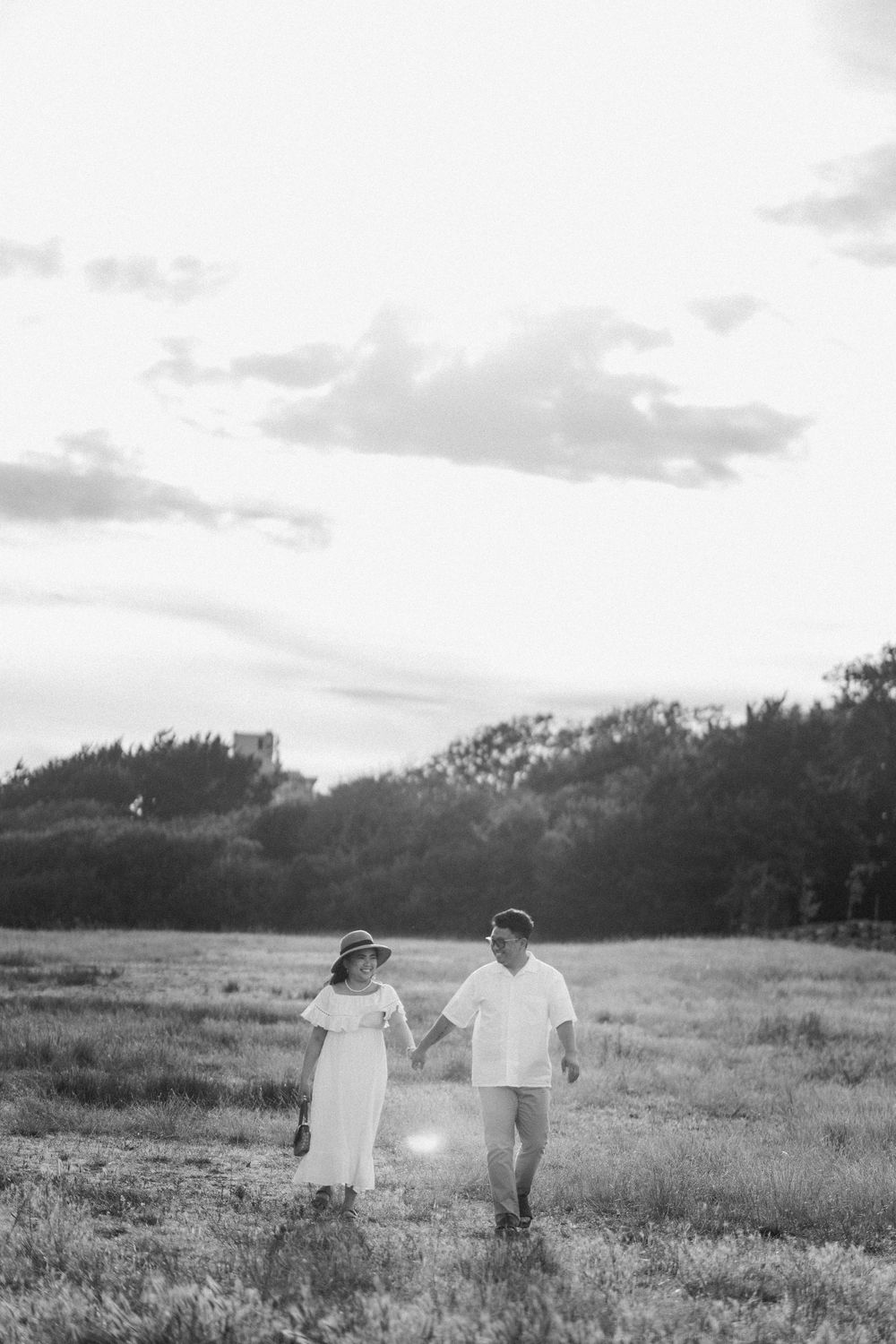 Black and white photo of couple holding hands walking through open field, woman in hat and white dress, romantic countryside.