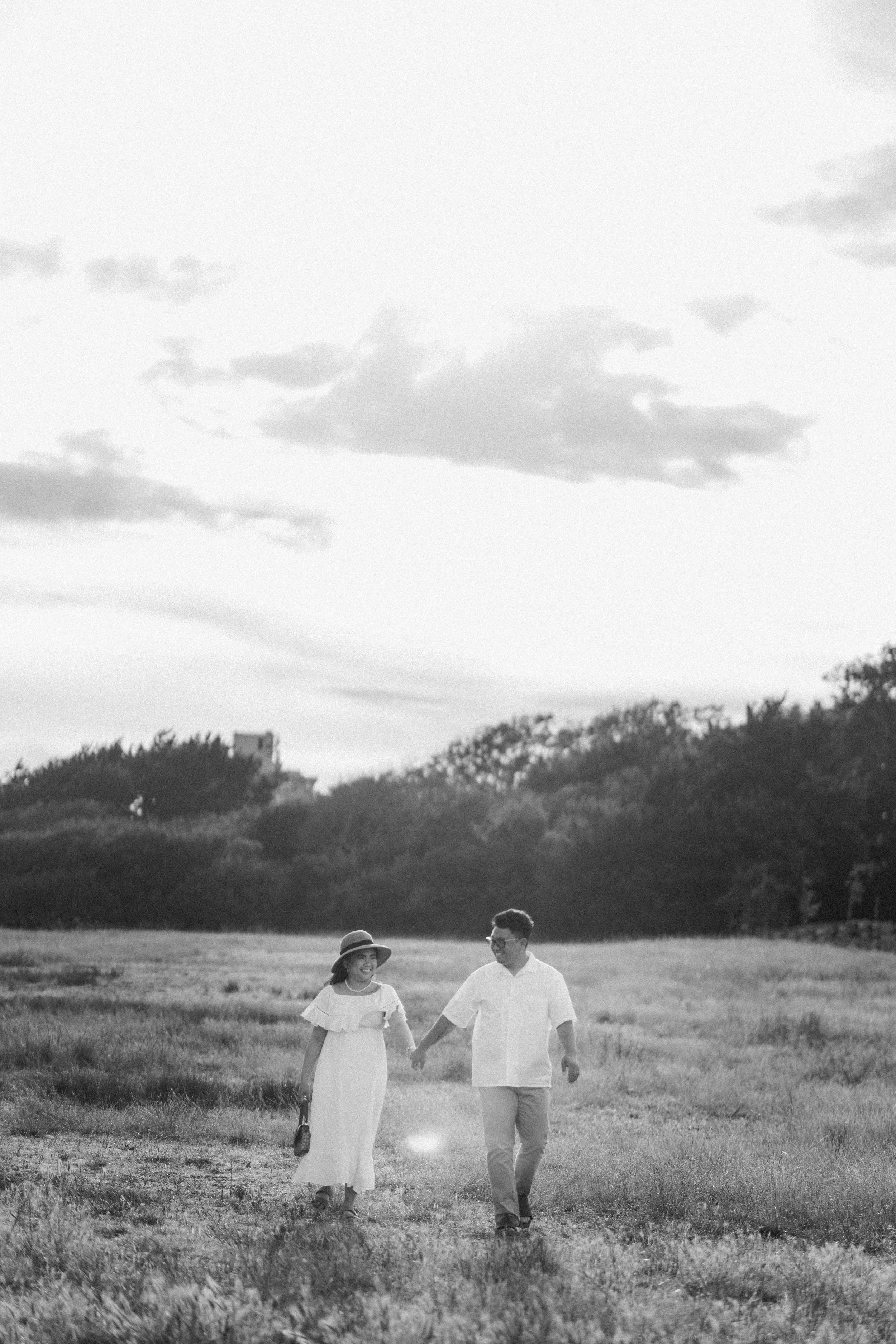 Black and white photo of couple holding hands walking through open field, woman in hat and white dress, romantic countryside.