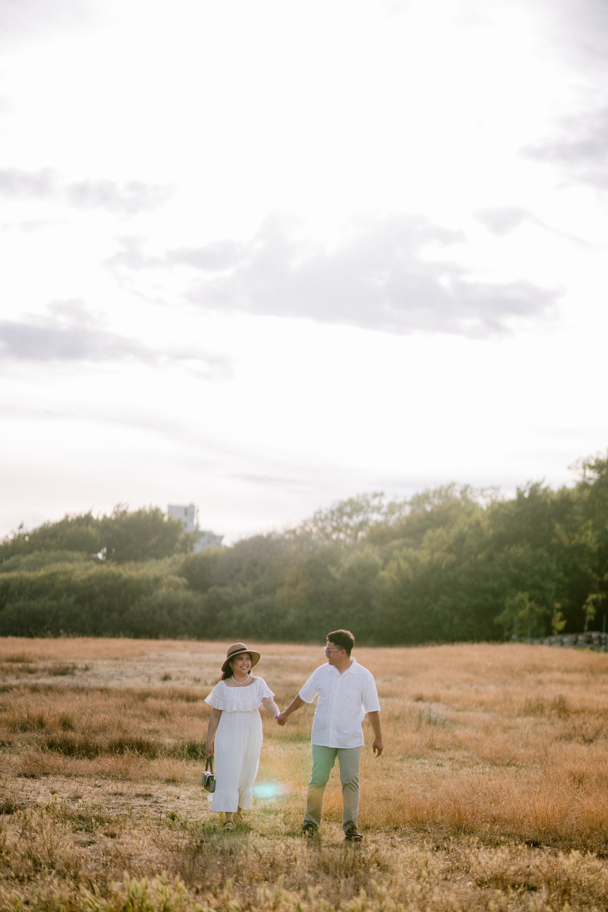 Couple holding hands in a sunlit golden field, woman in white off-shoulder dress, man in light green pants.