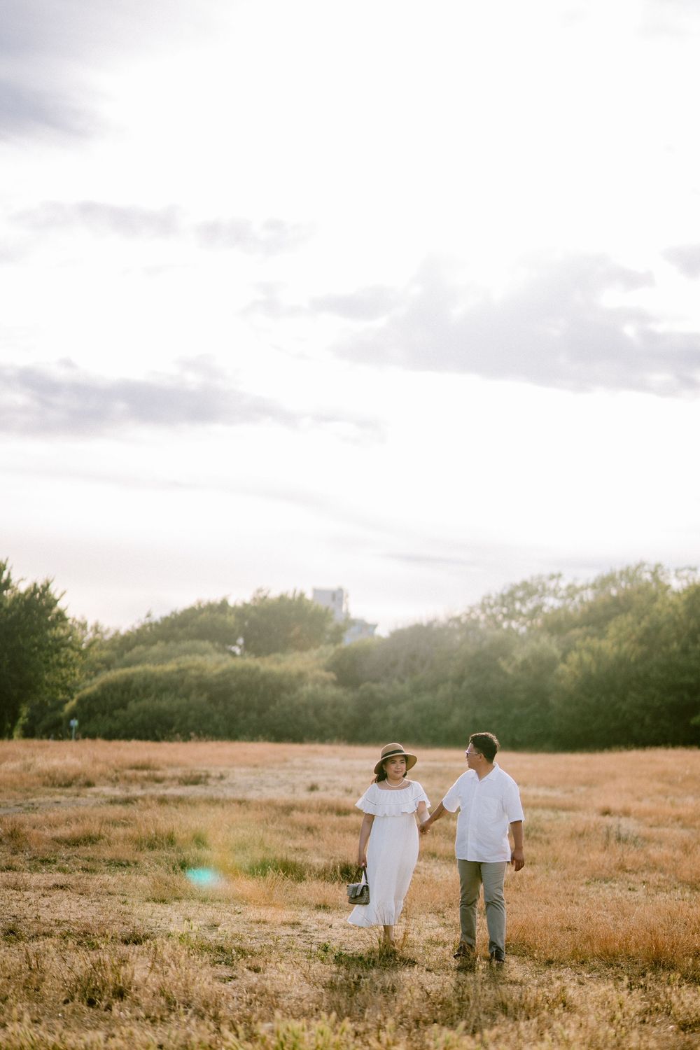Couple in white outfits hold hands while walking through a golden field with trees and cloudy sky backdrop.