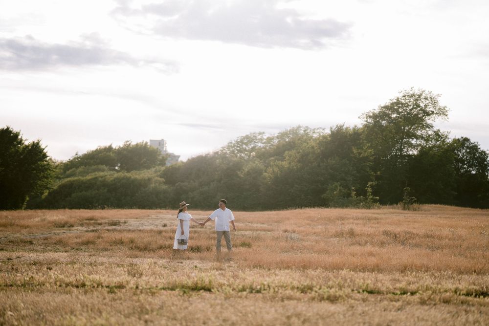 Couple holding hands in a golden field at sunset with lush green trees in the background.