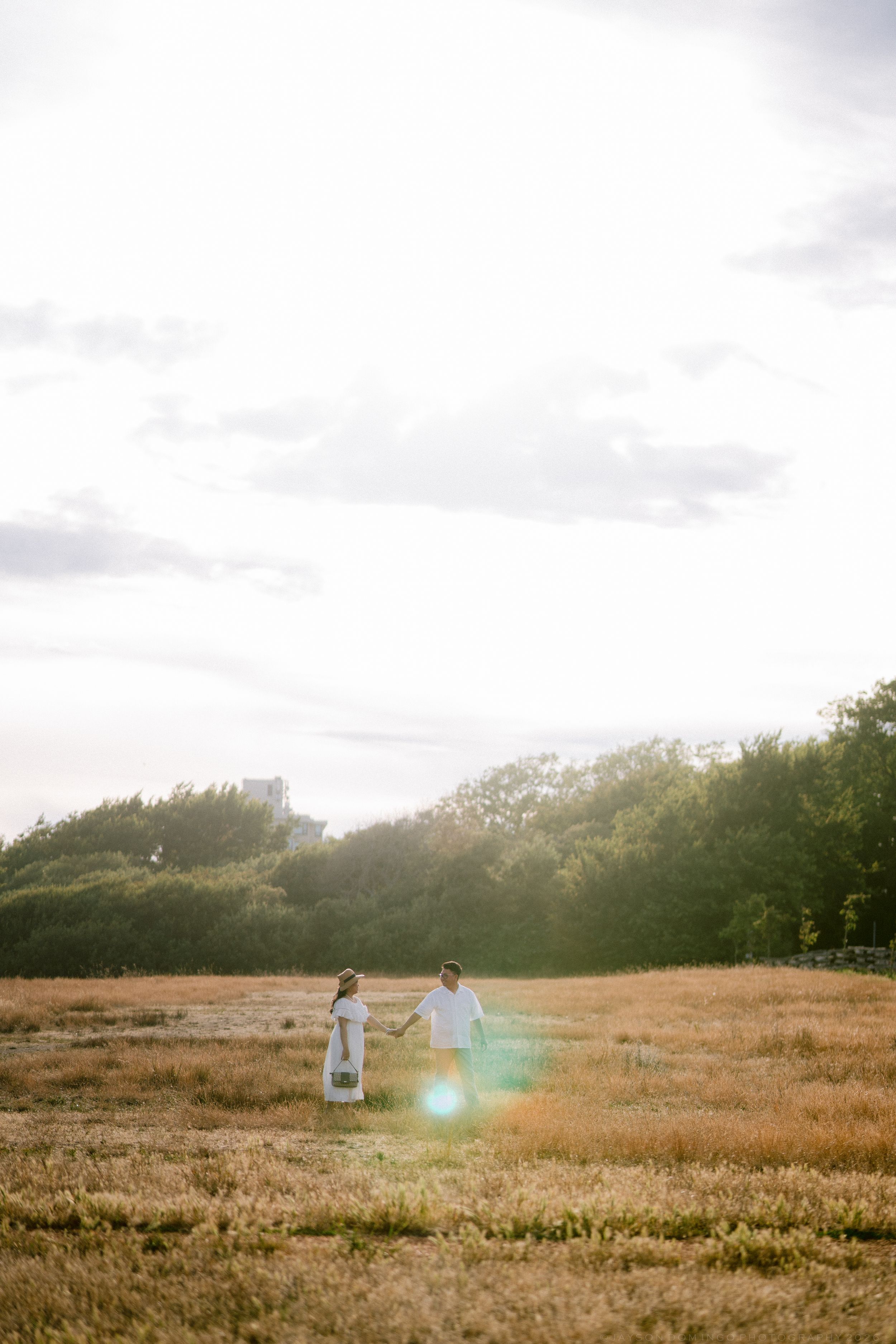Couple holding hands in golden field with sunflare and green trees at sunset.