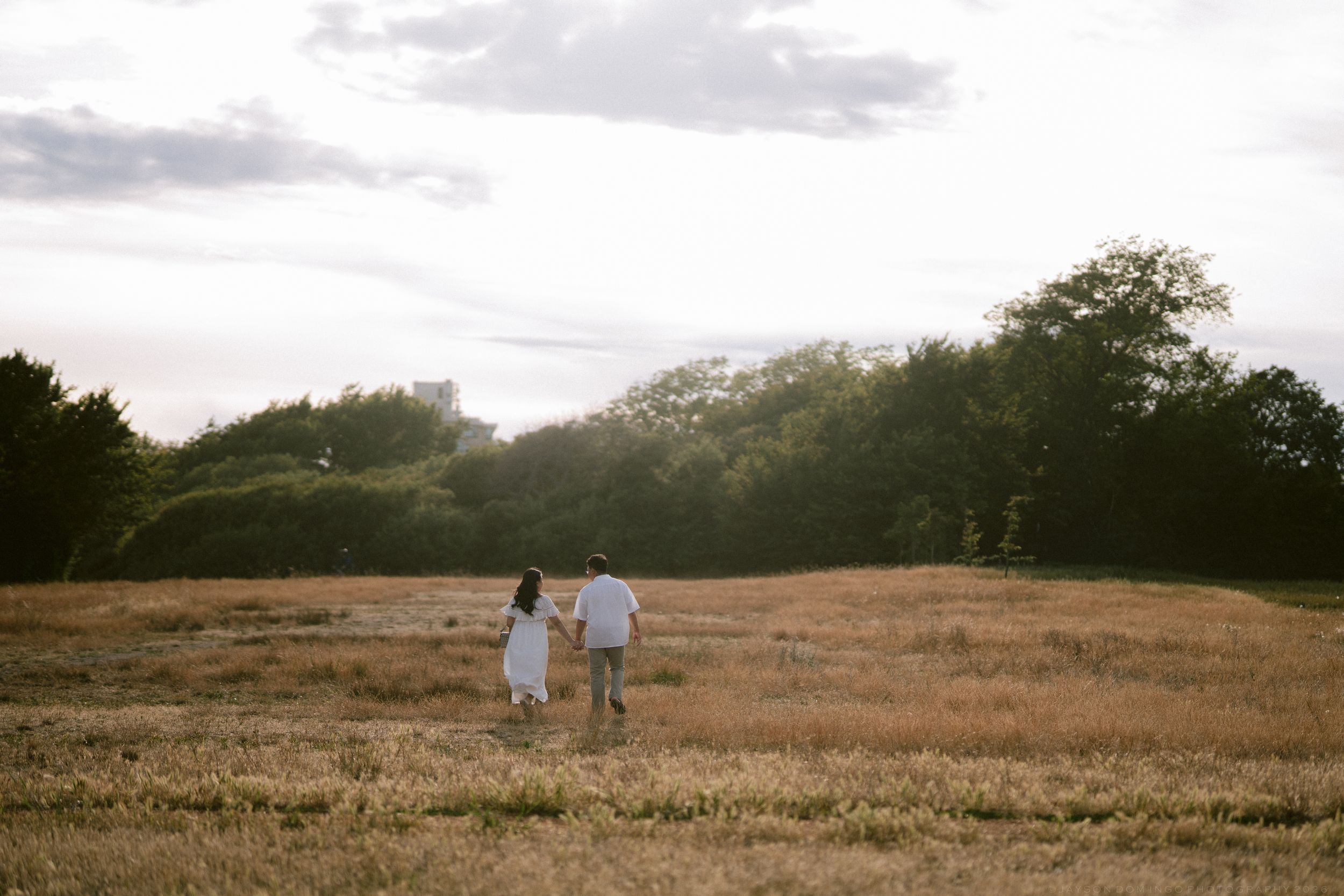 Couple walking hand in hand through golden meadow toward sunlit trees.