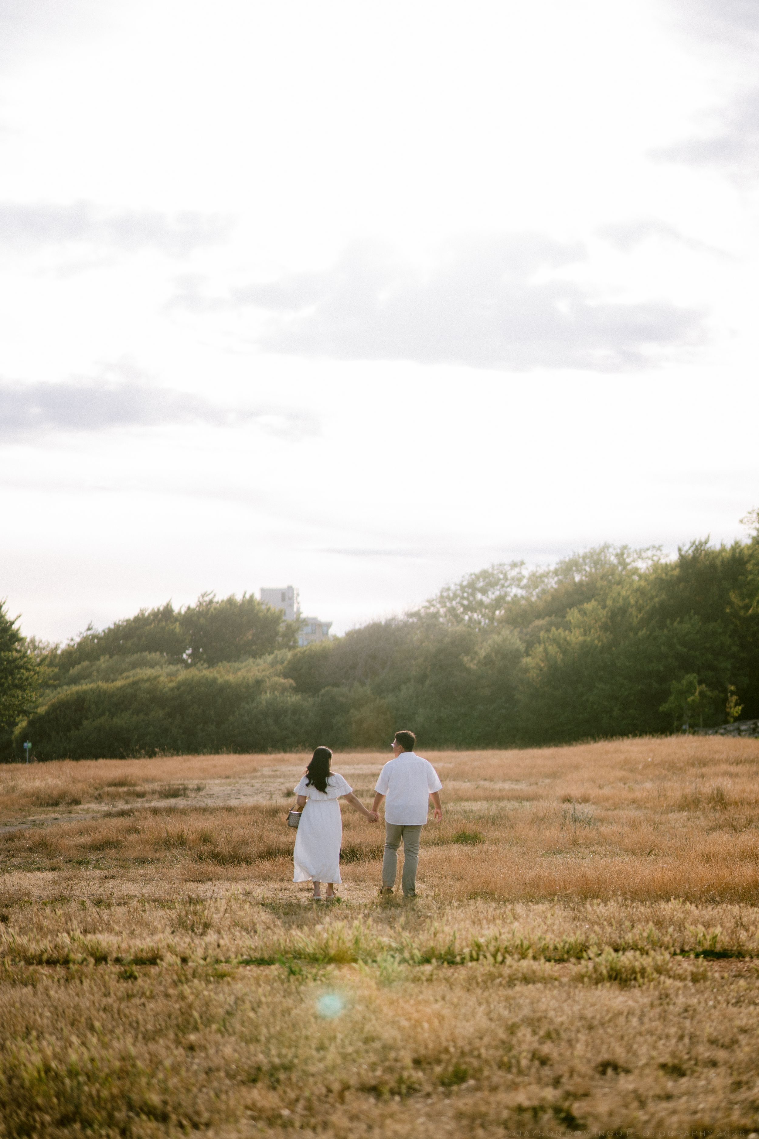 Couple walking hand in hand through a golden field at sunset, viewed from behind, surrounded by lush trees.