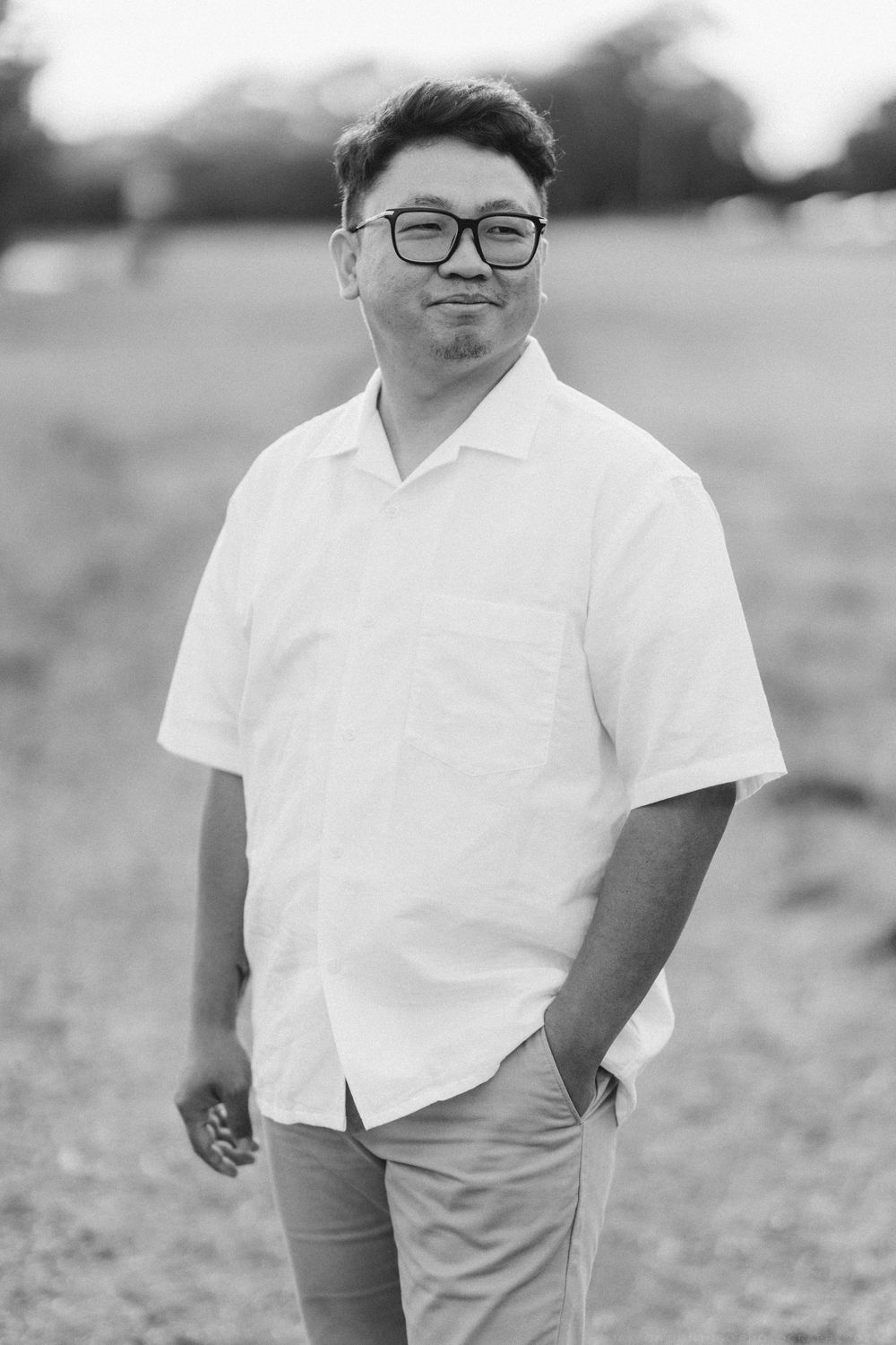 Black and white portrait of Asian man in white shirt and glasses in field.