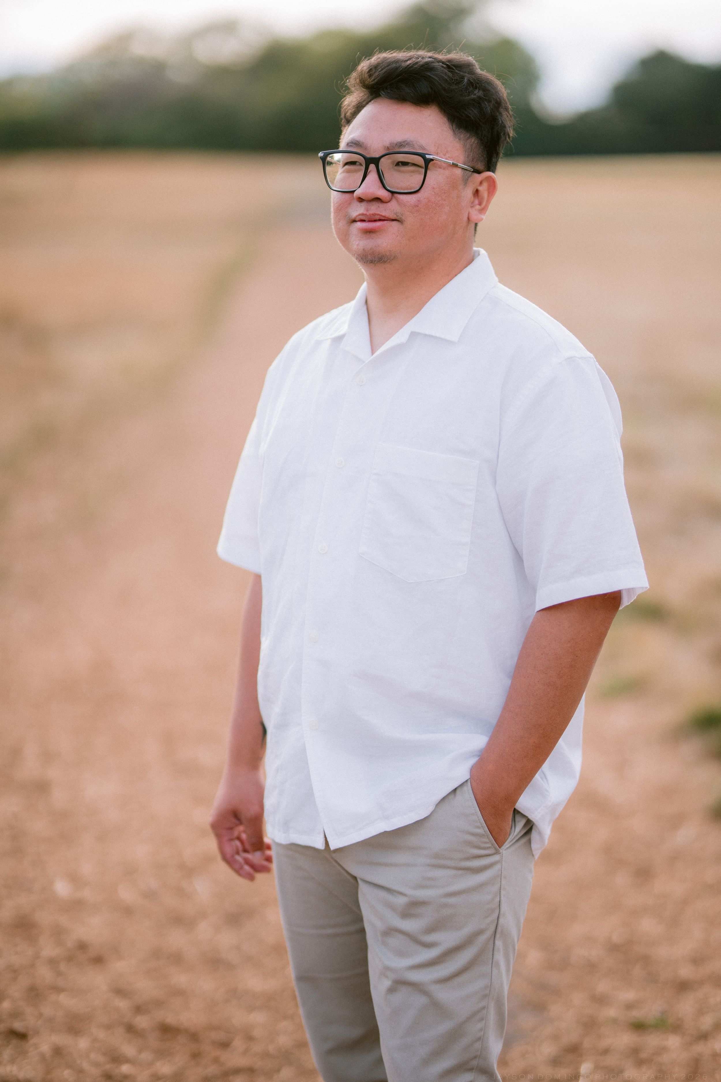 Asian man in white shirt and gray pants standing in harvested field wearing black glasses outdoors.