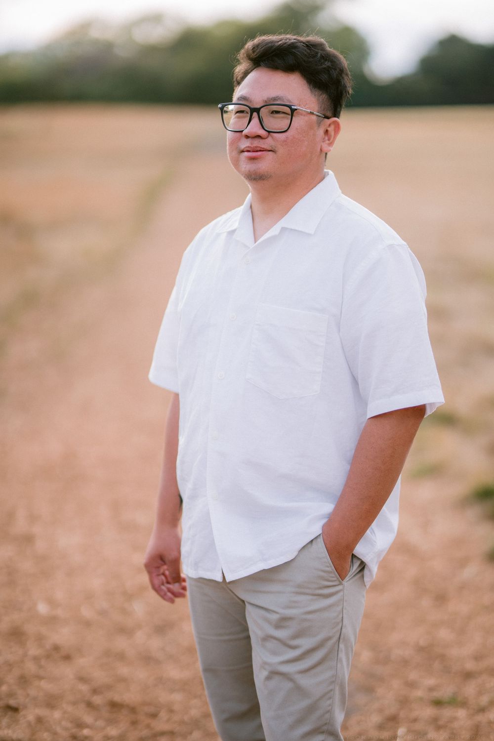 Asian man in white shirt and gray pants standing in harvested field wearing black glasses outdoors.