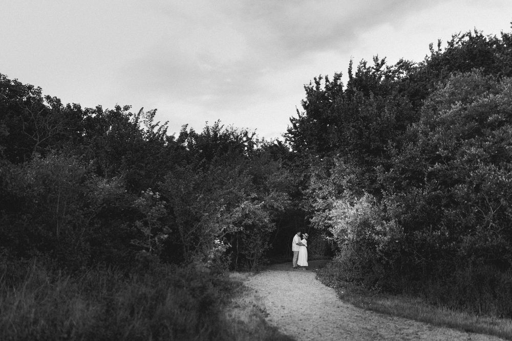 Black and white photo of a couple standing on a tree-lined path in a lush garden setting.