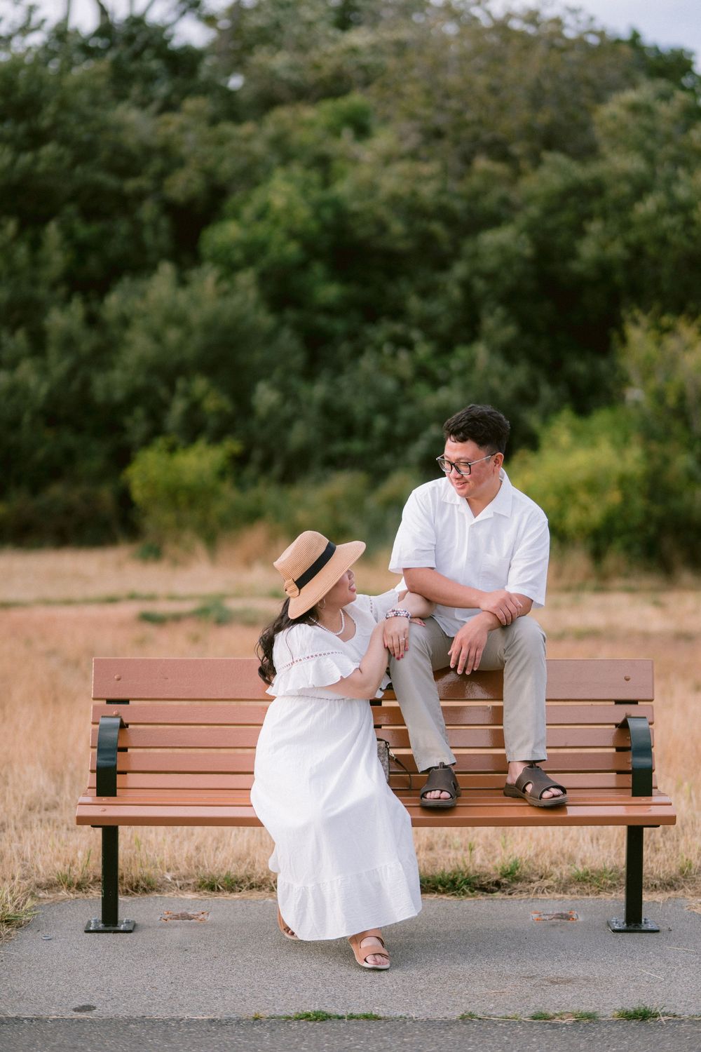 Couple in white outfits sitting on wooden bench in golden field at sunset.
