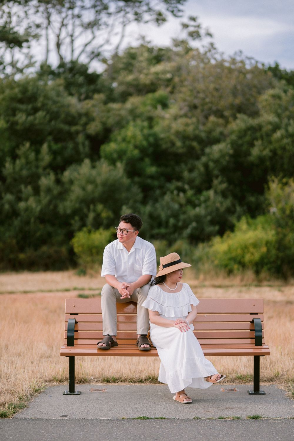 Couple in white outfits sitting on a park bench outdoors, woman wearing straw hat, lush green trees in background.