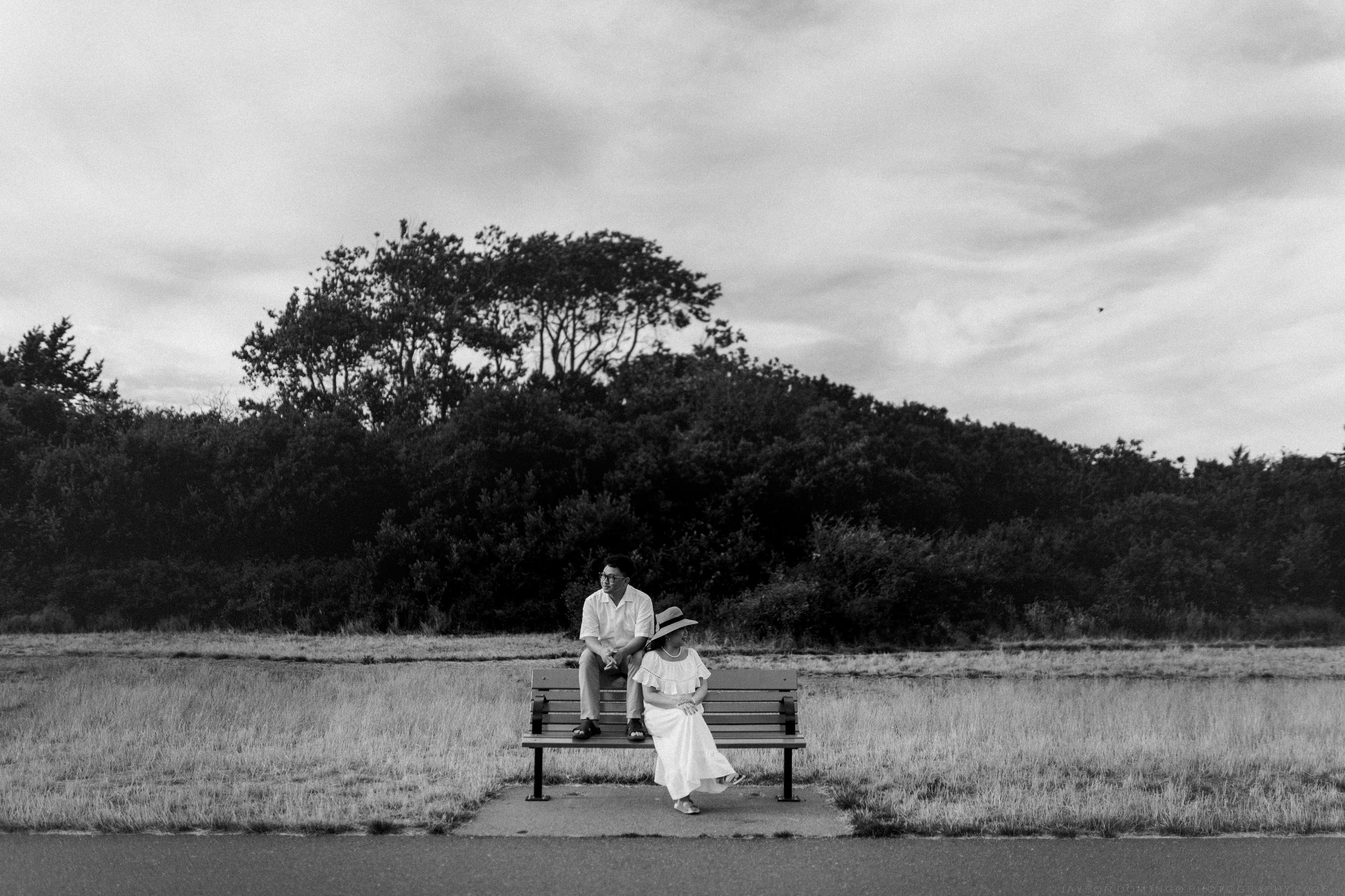 Black and white photo of couple sitting on bench near field and trees.