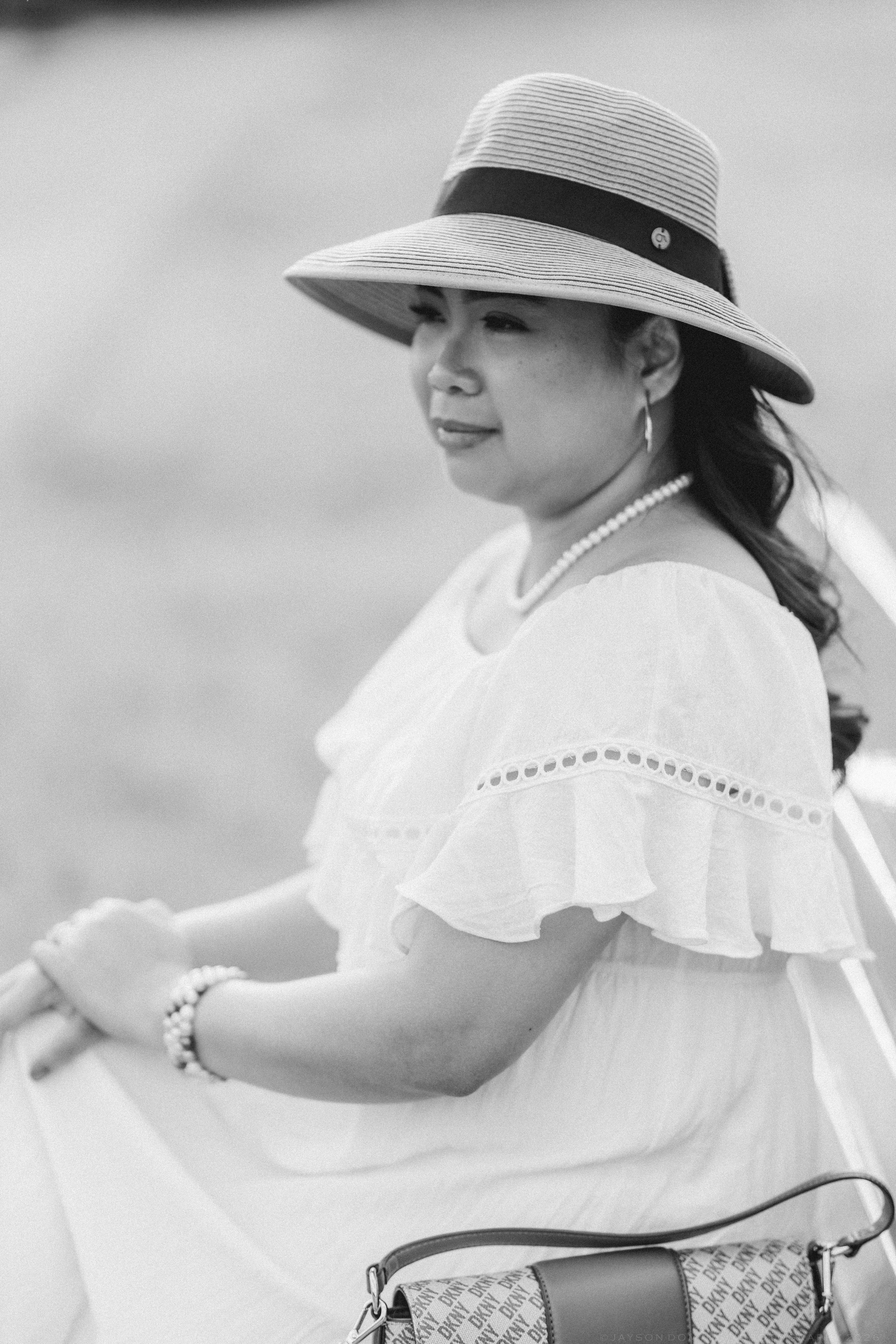 Black and white portrait of a woman in a wide-brim straw hat, white ruffled dress, and pearl necklace outdoors.
