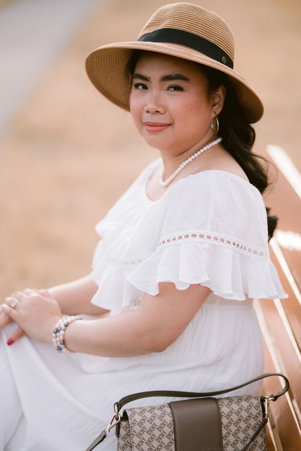 Smiling Asian woman in white off-shoulder dress and straw hat sitting on bench with designer handbag.