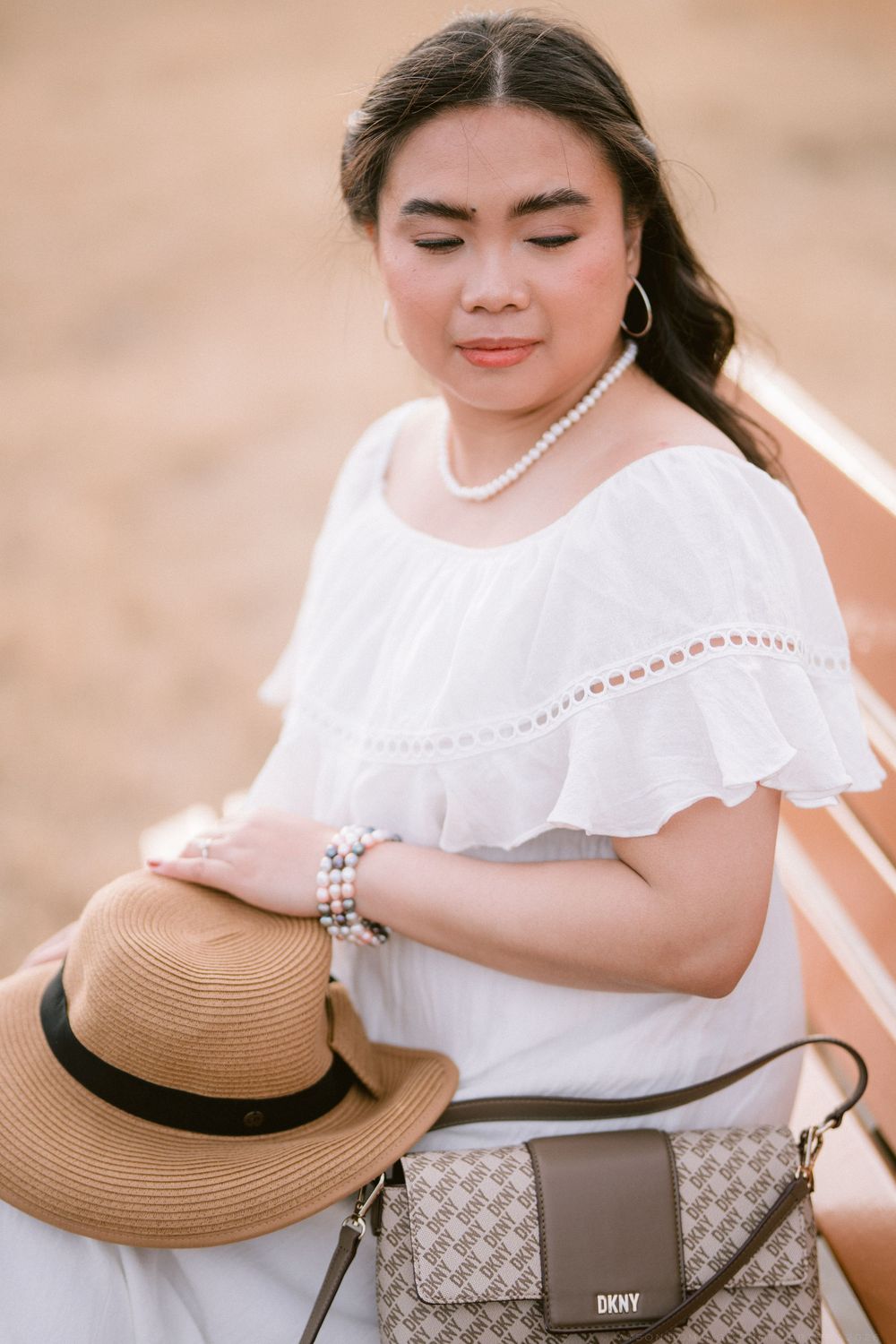 Woman in white off-shoulder dress holding straw hat with DKNY bag on bench.