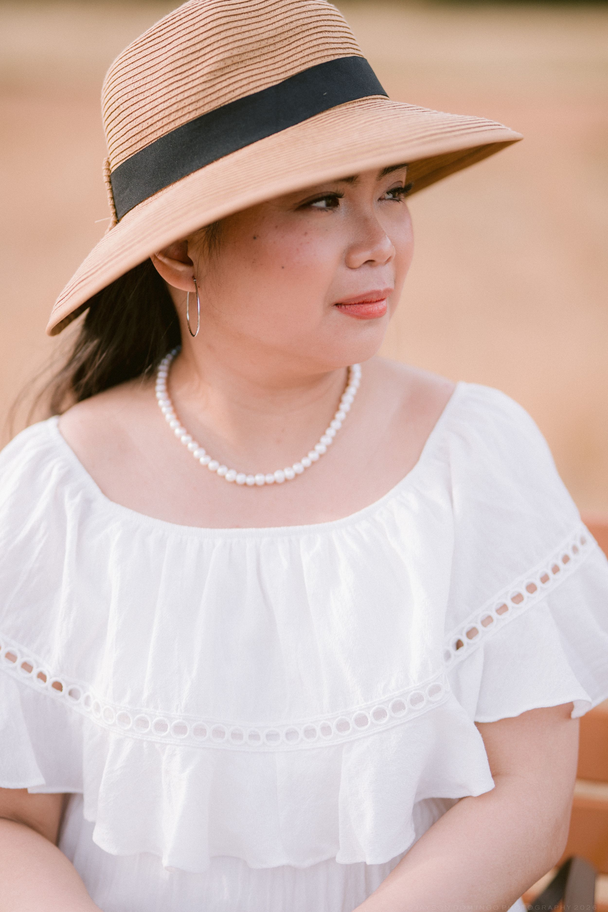 Close-up portrait of a woman wearing a striped sun hat with black ribbon, pearl necklace, and white off-shoulder dress.