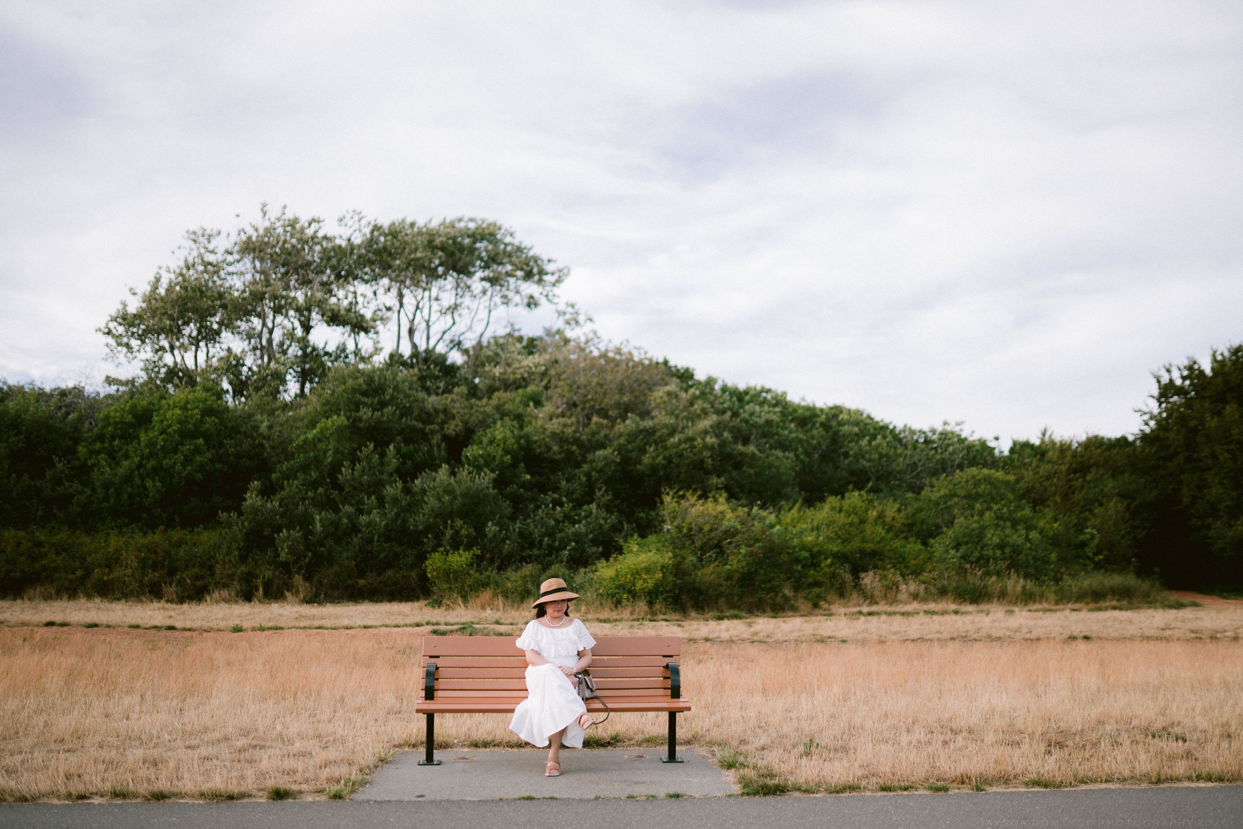 Woman in white dress and straw hat sitting on wooden bench surrounded by lush greenery and dry grass field.