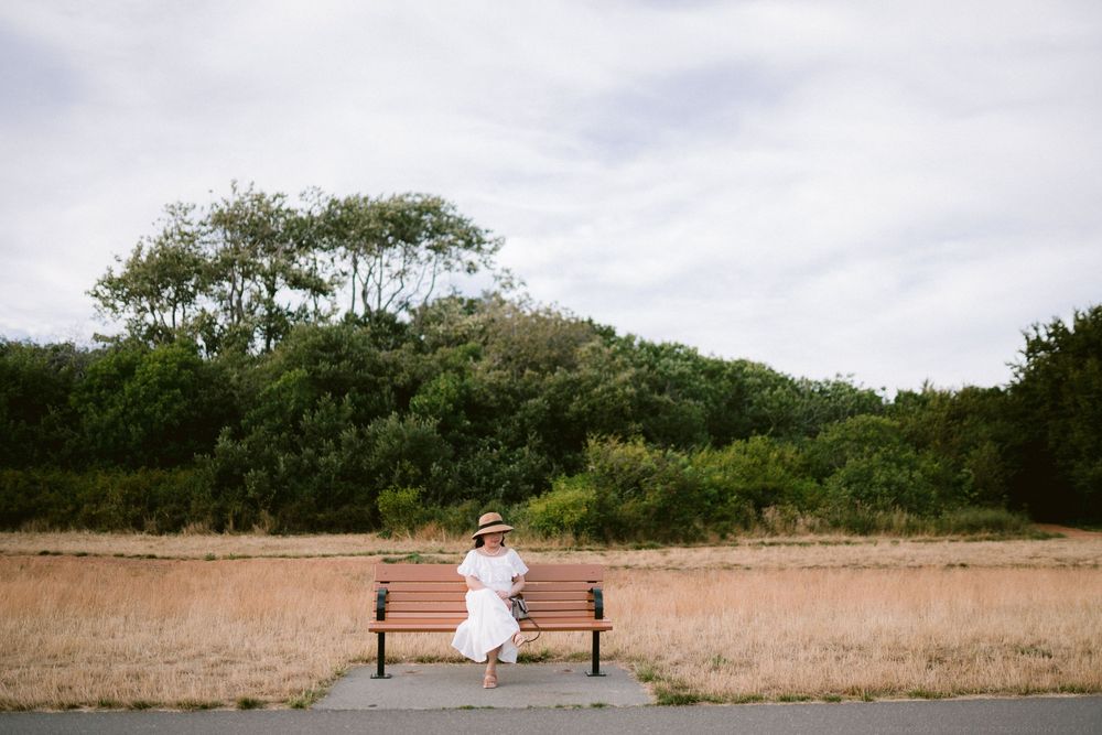 Woman in white dress and straw hat sitting on wooden bench surrounded by lush greenery and dry grass field.