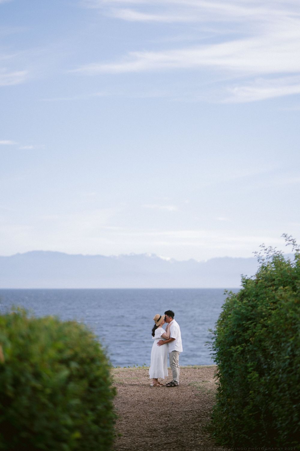 Couple embracing near the ocean shore, surrounded by lush green hedges with mountains visible in the background.