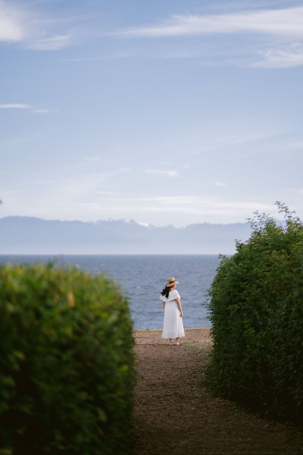 Woman in white dress and straw hat standing between green hedges gazing at ocean with snow-capped mountains in distance.