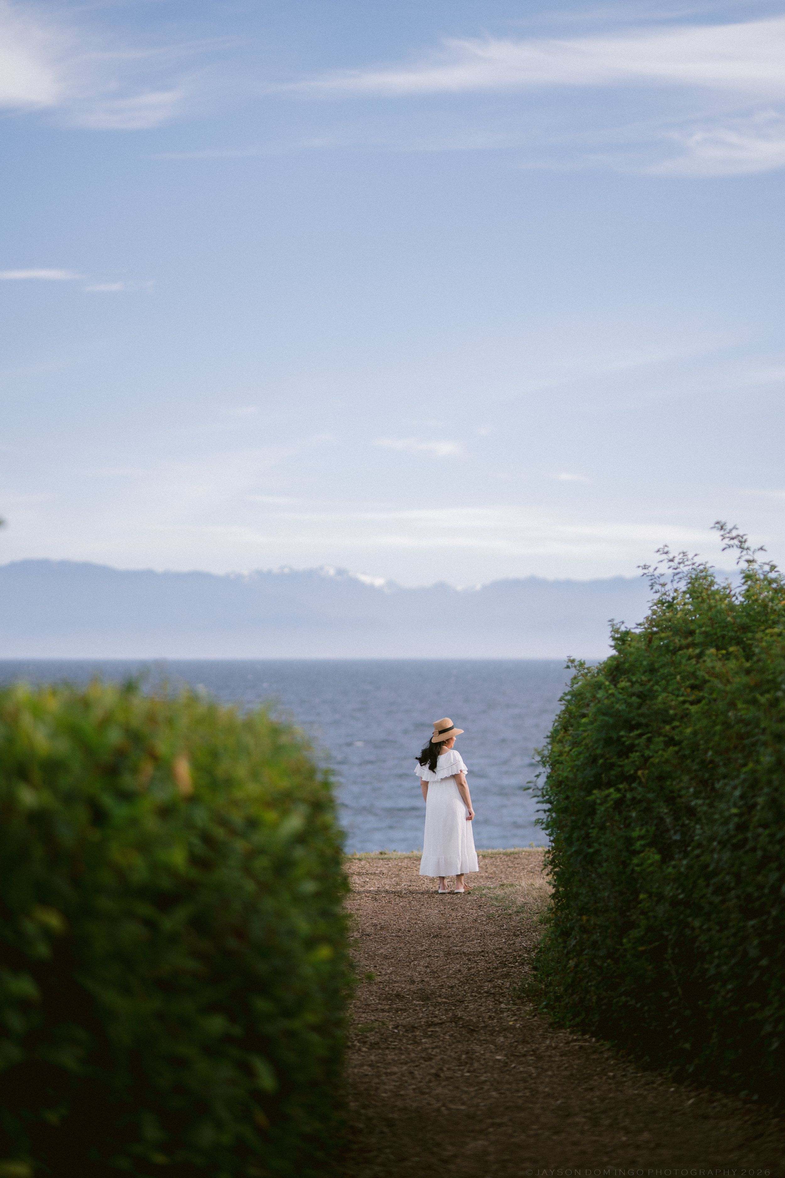 Woman in white dress and straw hat standing between green hedges gazing at ocean with snow-capped mountains in distance.