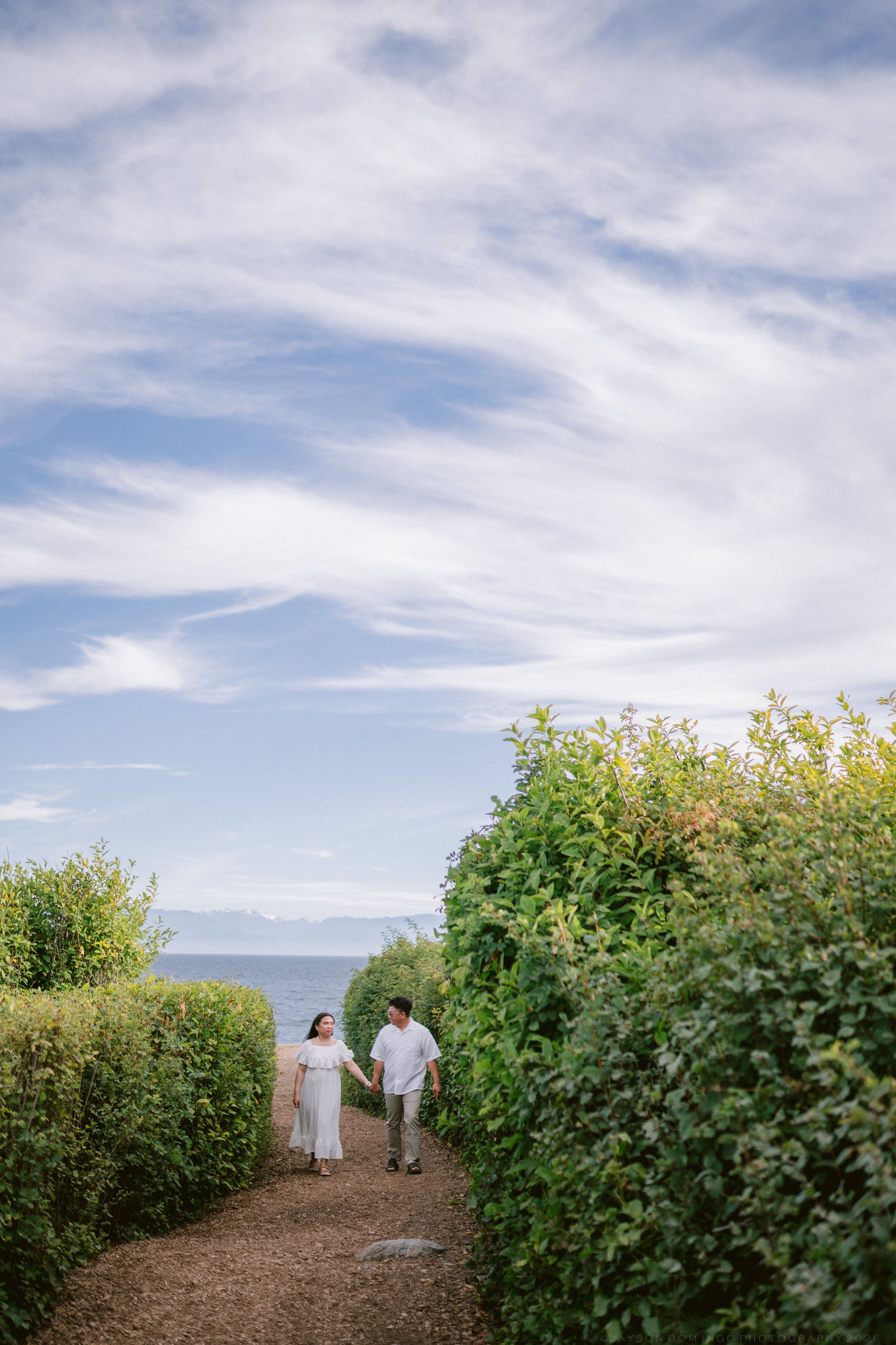 Couple in white holding hands walking on path between green hedges near ocean.