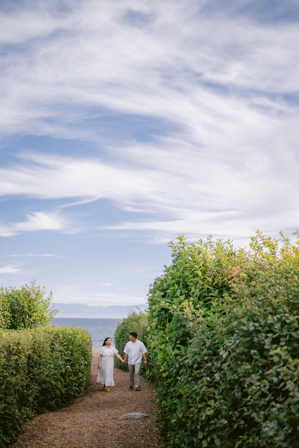 Couple in white holding hands walking on path between green hedges near ocean.