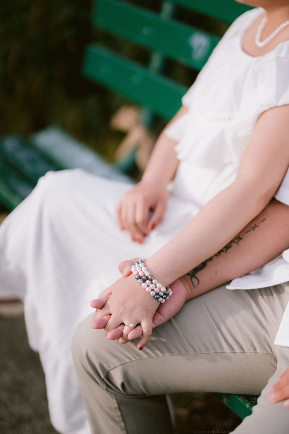 Close-up of couple holding hands showing pearl bracelet and tattoo on bench.