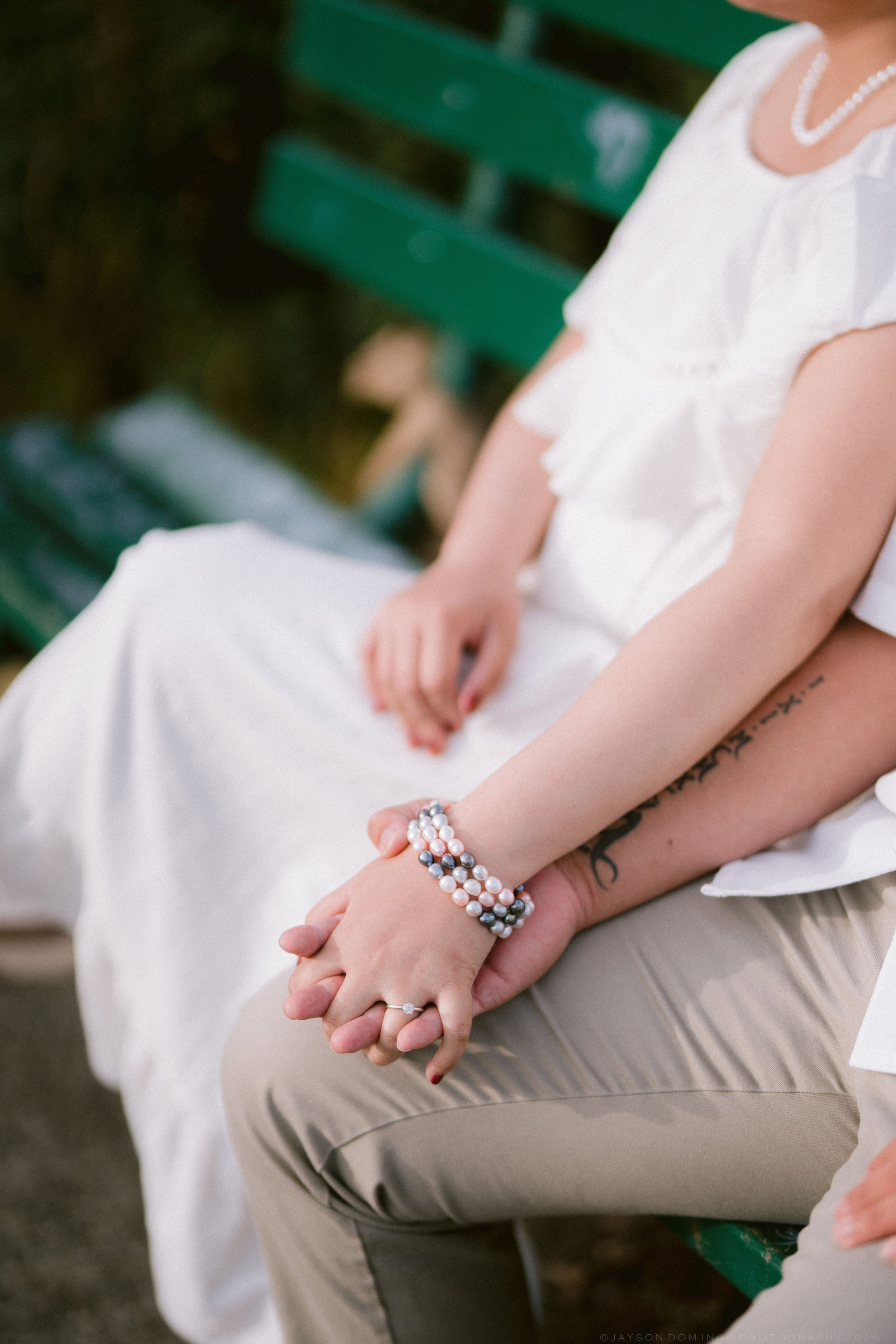 Close-up of couple holding hands showing pearl bracelet and tattoo on bench.