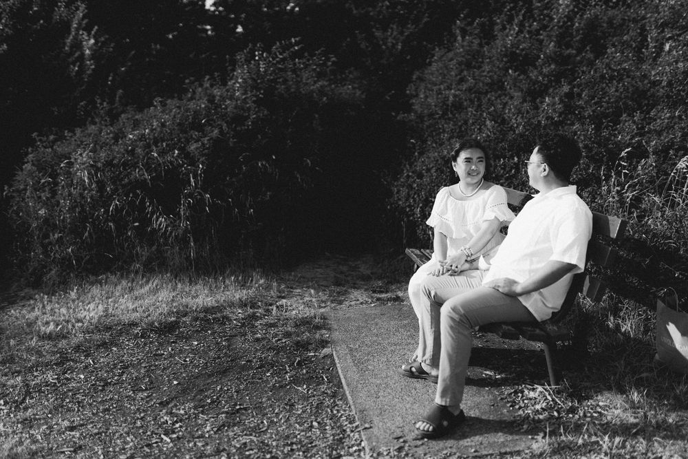 Two people sitting on a bench outdoors, engaged in conversation, surrounded by lush greenery in black and white.