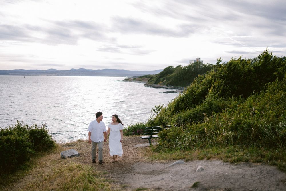 Couple walking hand in hand along scenic coastal path with ocean and mountains behind.