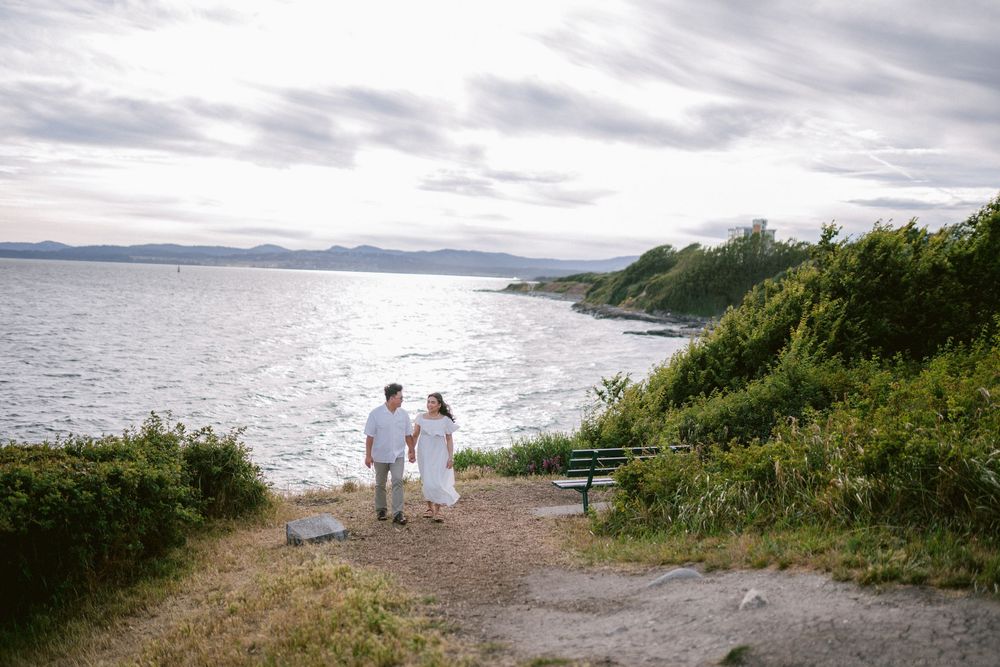 Couple in white outfits hold hands on a scenic coastal path with ocean views and lush greenery.